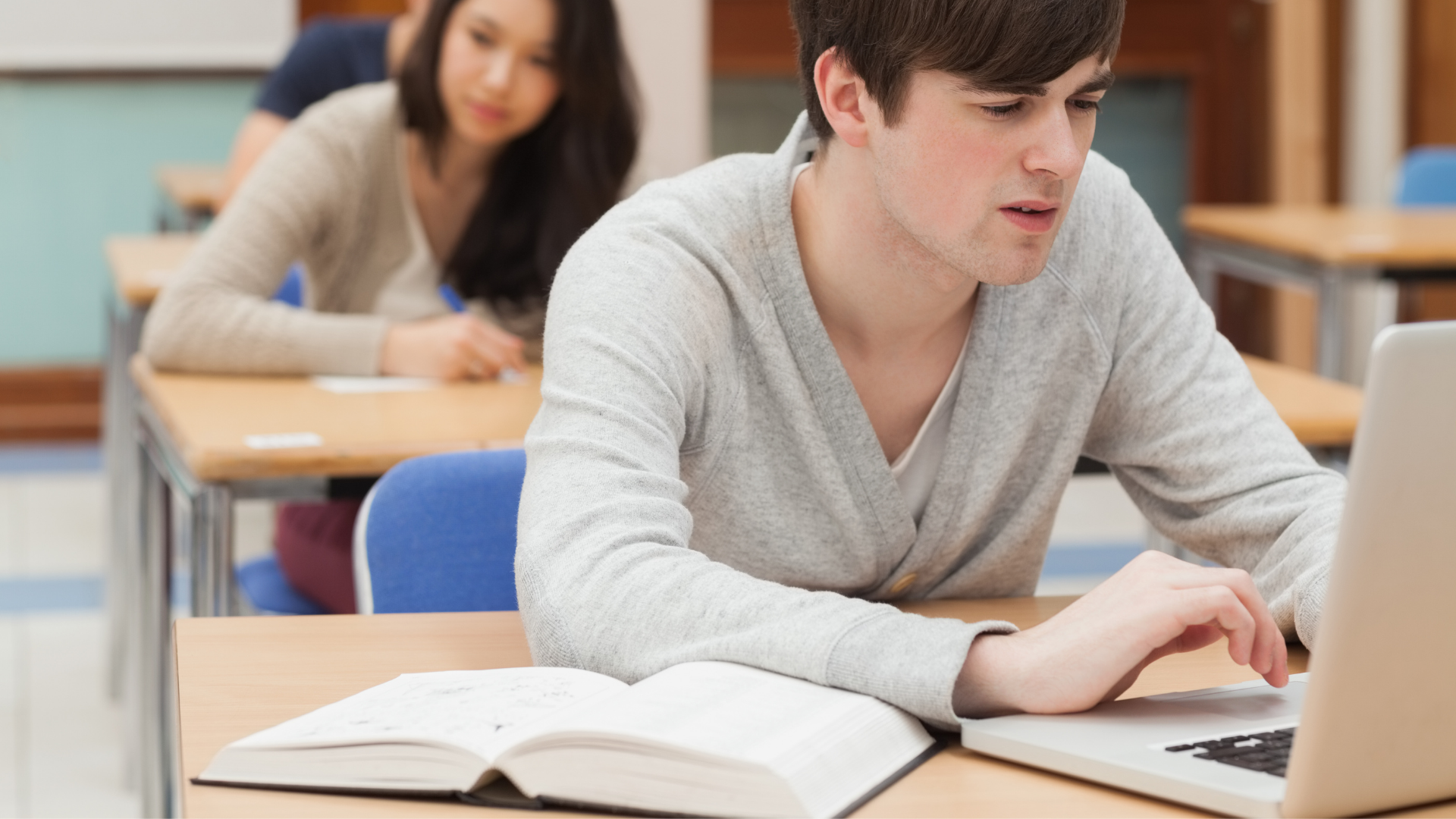student in grey jumper typing on laptop next to textbook